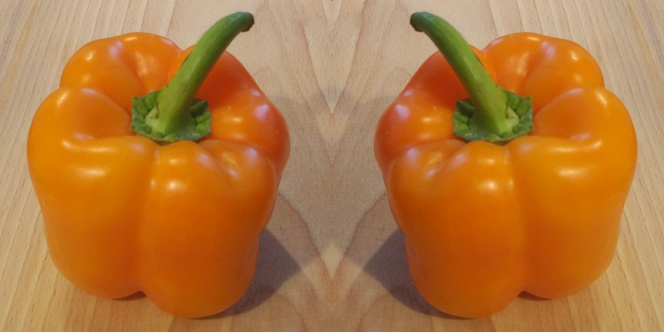 A tasty-looking orange bell pepper is sitting on a wooden cutting board in the left half of the image. The right half of the image is an exact copy of the left half, except horizontally flipped, giving the impression of a mirror plane at the centre.
